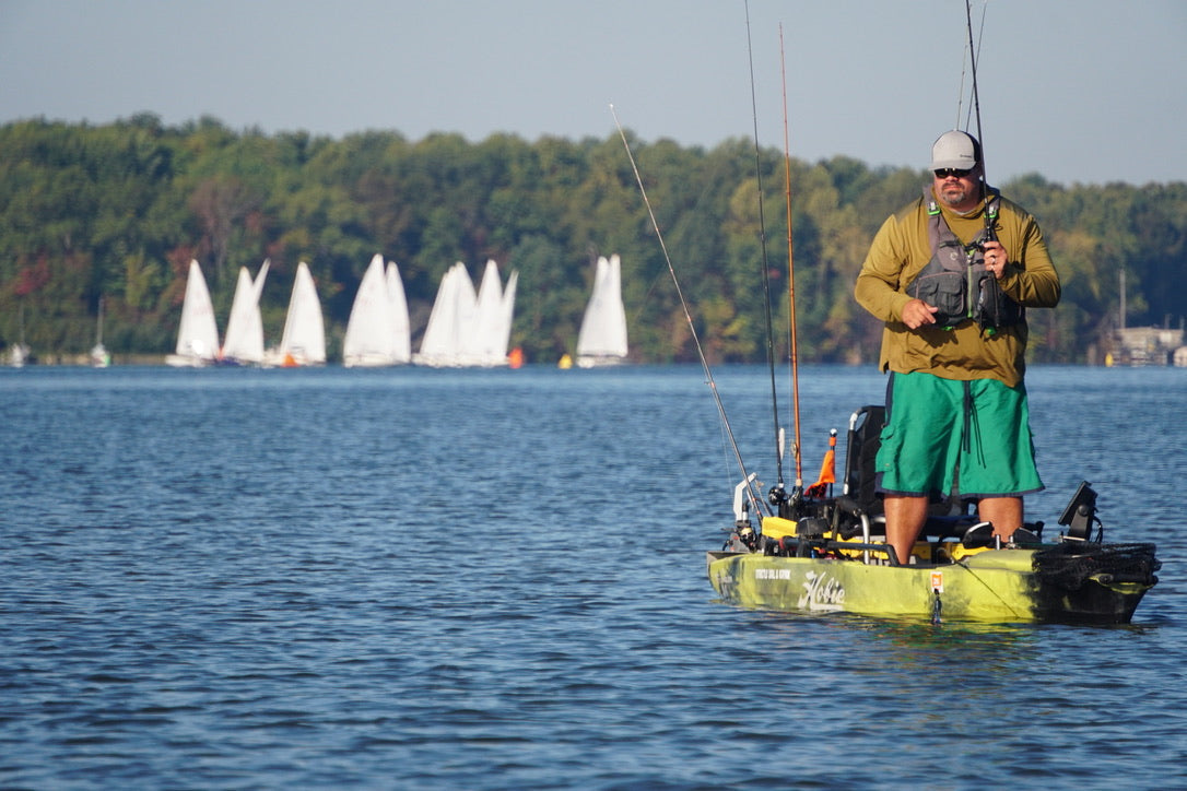 Angler standing in a fishing kayak with sailboats and a wooded shoreline in the background.