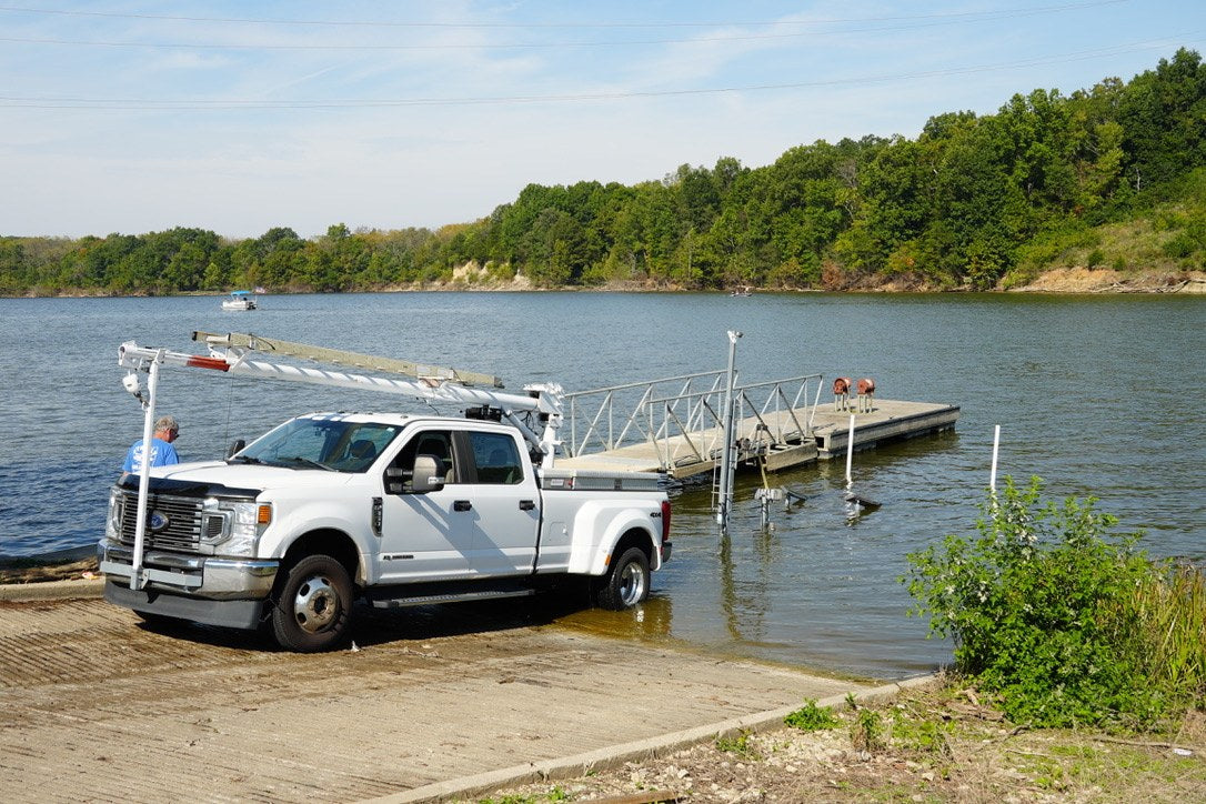 A white pickup beside a lakeside boat launch, with a dock and wooded shore beyond.