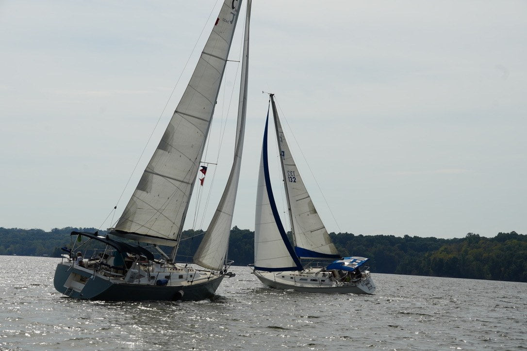 Two sailboats with white sails travel across a lake near a tree-lined shore.